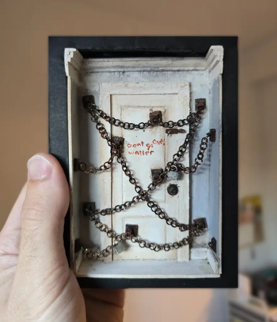 A diorama of Henry's door from Silent Hill 4. The door is locked with several rusted chains draping across the face of the door. Written on the door is a message in red ink: "don't go out!" signed by "Walter."