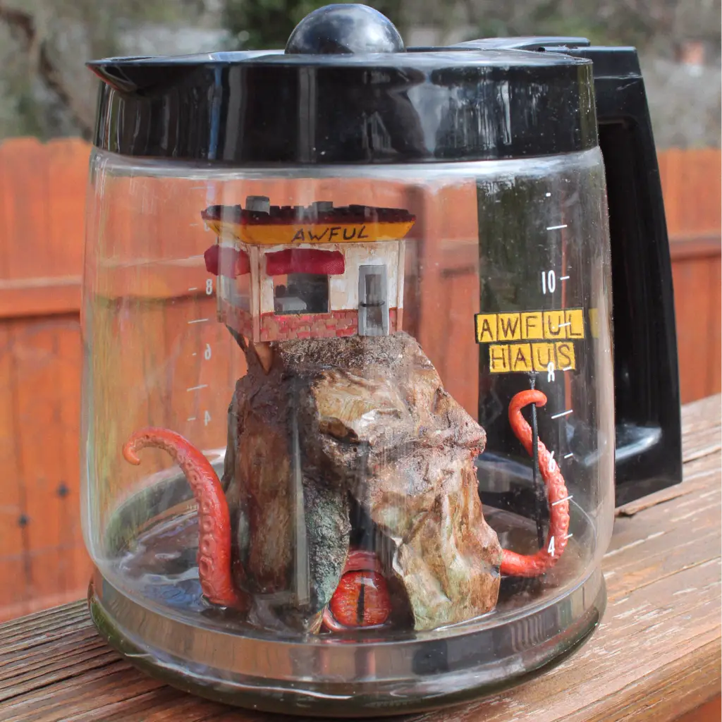 A diorama housed in a coffee pot. Inside is a diner, located on a treacherous cliff face. Emerging from the rocks below is an octopus-like monster, its eye visible from a crack in the cliff. One of its tentacles extends, holding a sign that says "AWFUL HAUS."